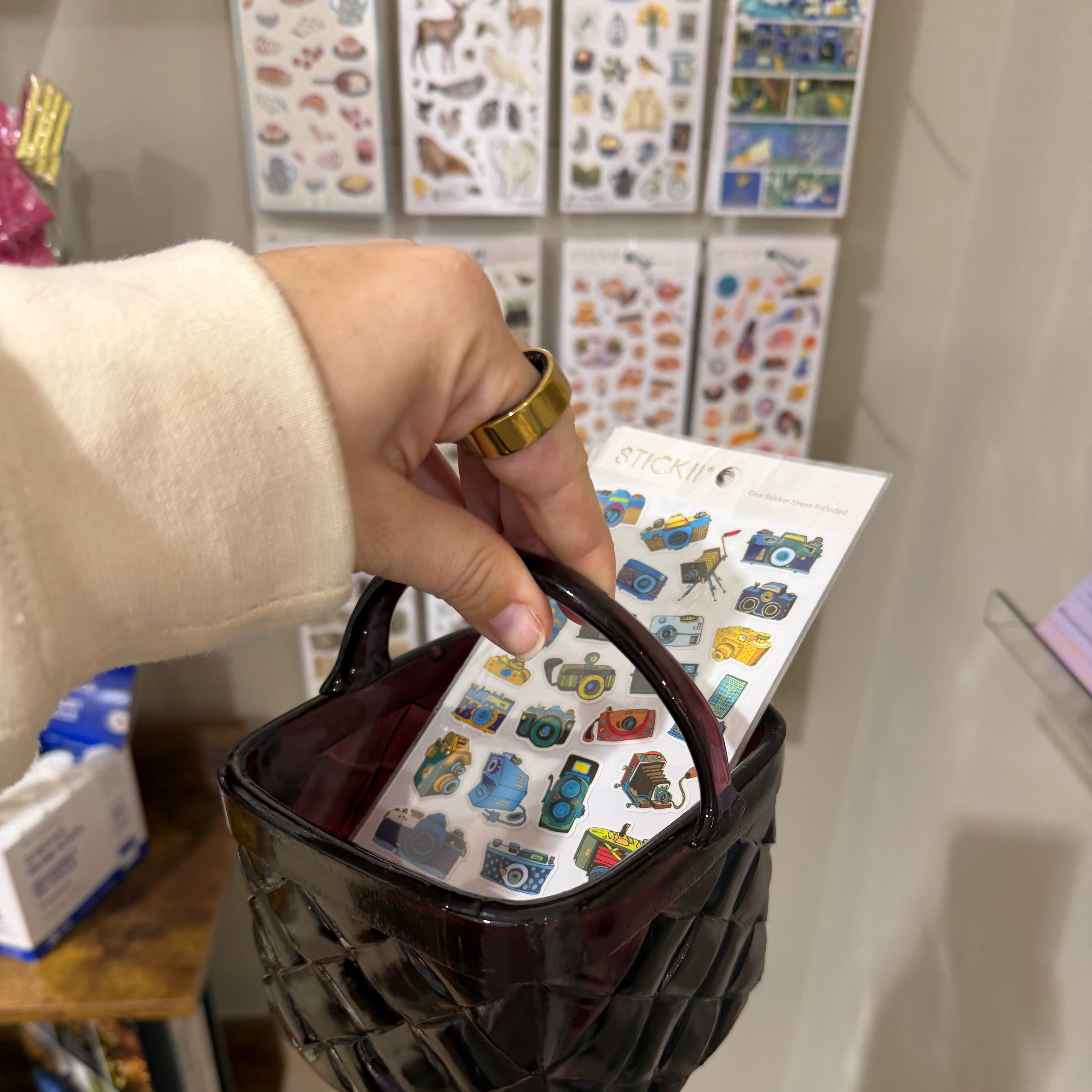 Hand holding a small black bag with colorful stickers against a wall of sticker sheets.